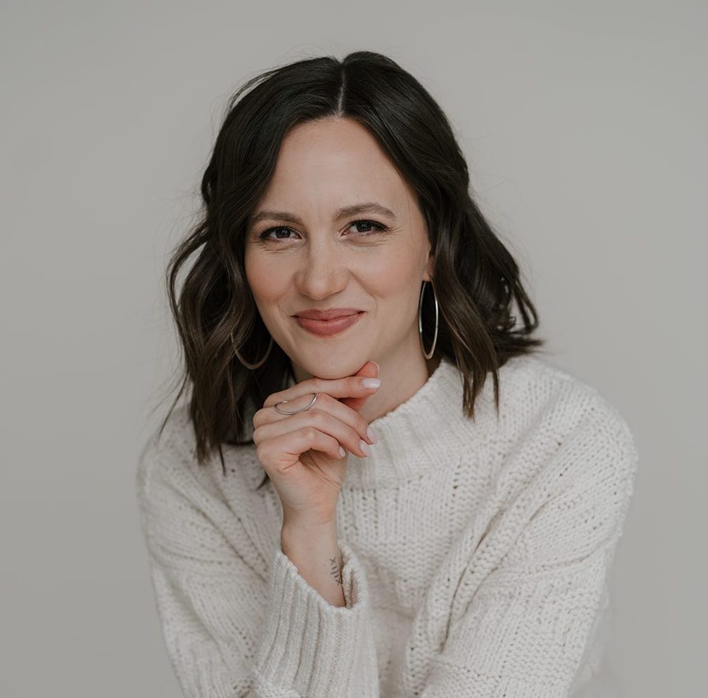 Chantal is a woman with shoulder-length dark brown hair, brown eyes and light skin. She is sitting in a relaxed pose, wearing a white knit sweater and blue jeans. She is smiling gently and resting her chin on her hand. The background is a neutral beige color.