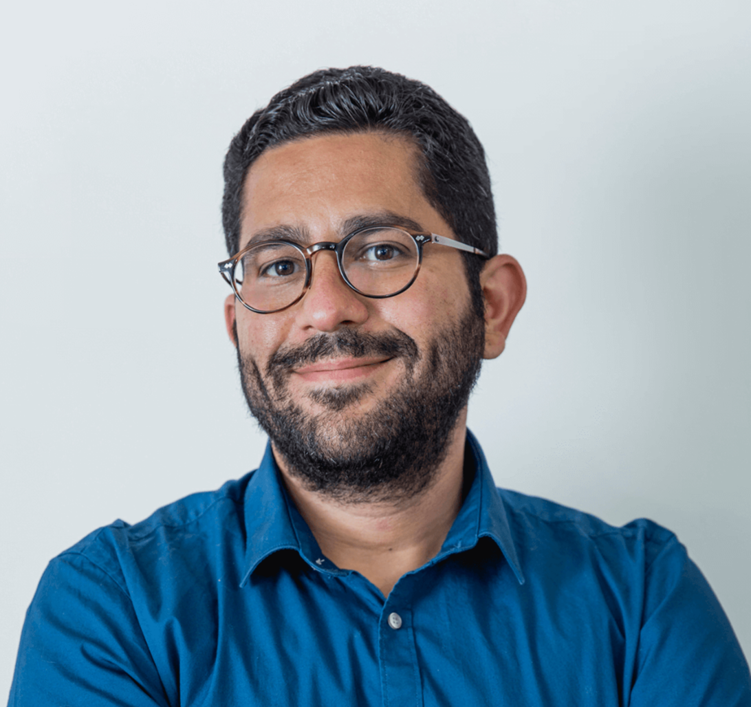 Mostafa is a caucasian man with black beard and is wearing glasses. He is smiling at the camera. He is wearing a blue shirt.