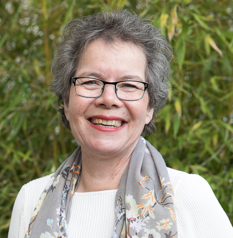 Tessa is a white woman with short greyish-brown hair and green-blue eyes. She is wearing a white sweater with a grey scarf in front of green bamboo leaves.