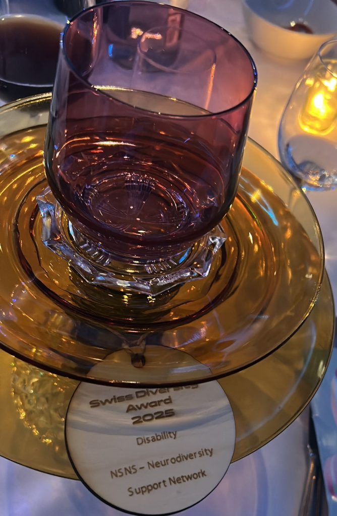 This image depicts a glass trophy consisting of two stacked platforms and a glass cup on top. The wooden plaque reads: Swiss Diversity Award 2025 Disability NSNS - Neurodiversity Support Network Switzerland