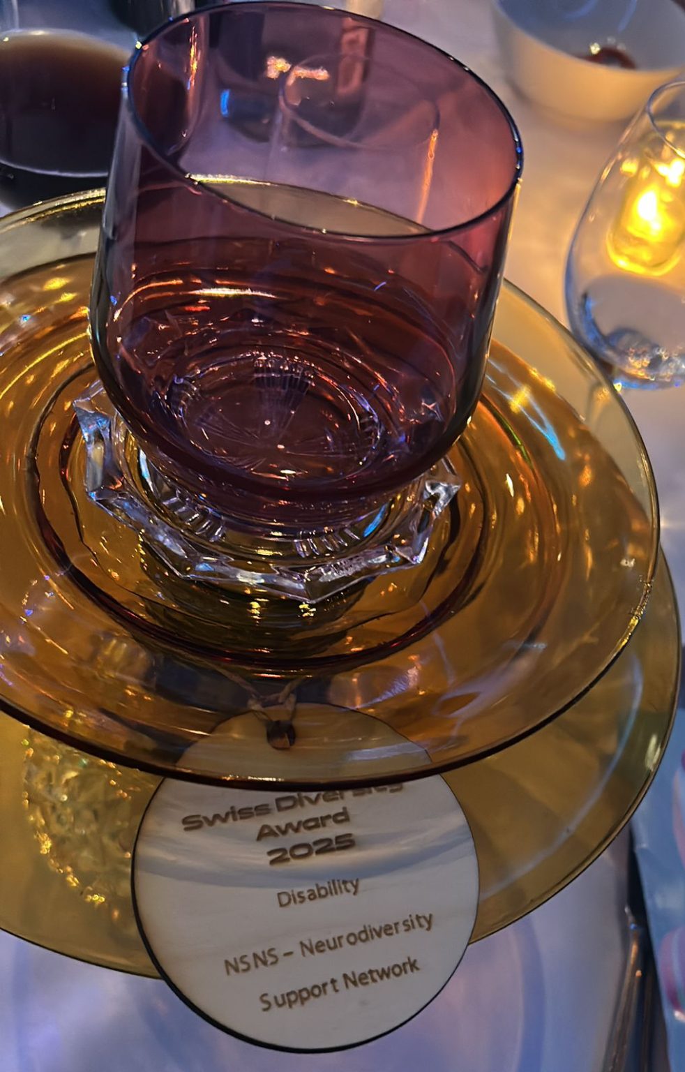 The image depicts a glass trophy with two stacked circular platforms and a glass cup sitting on top. The circular wooden plaque reads: Swiss Diversity Award 2025 Disability NSNS - Neurodiversity Support Network Switzerland"