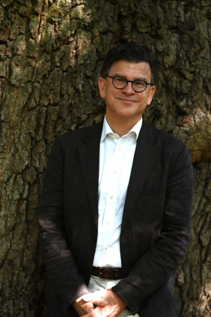 Jens is a white man with dark hair and glasses. He is wearing a white chemise with a black coat. He is standing against a tree and is facing the camera.