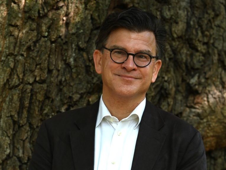 Jens is a white man with dark hair and glasses. He is wearing a white chemise with a black coat. He is standing against a tree and is facing the camera.