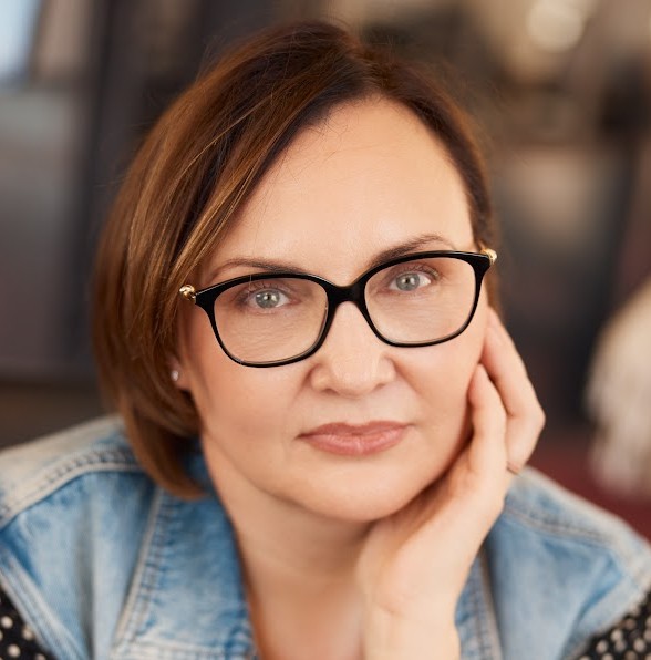 Lucy is a white woman with short brown hair and glasses. She is wearing a blue shirt and is resting her chin on her hand. She is looking into the camera