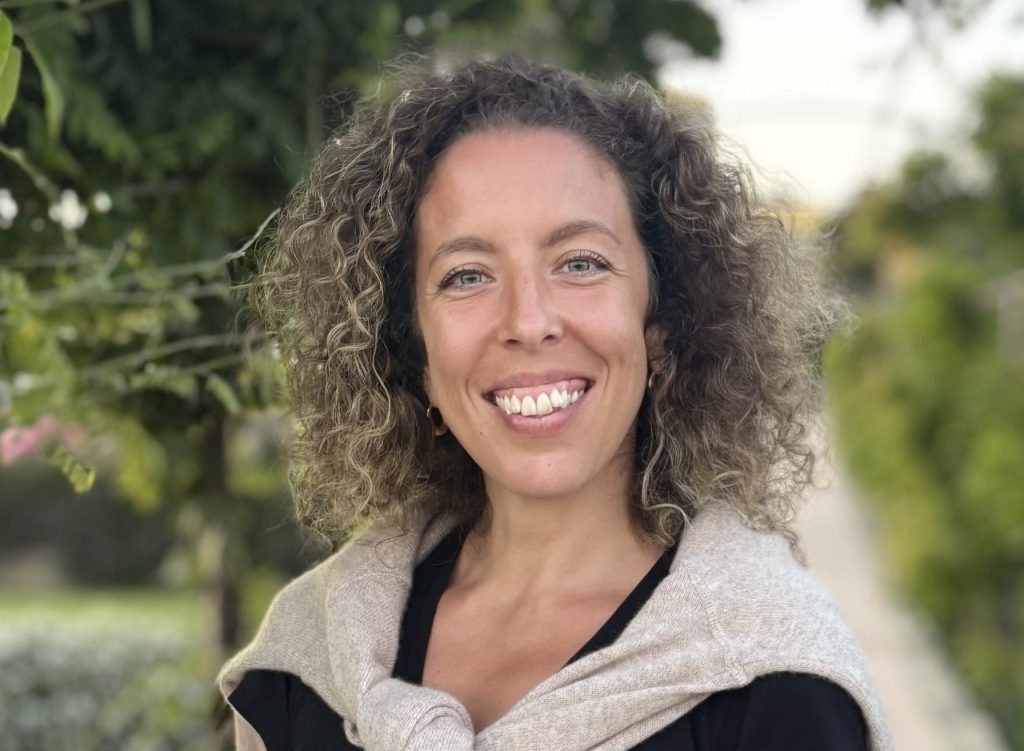 Laura is a fair skinned woman with curly brown hair. She is wearing a black shirt with a tan sweater and is smiling into the camera.