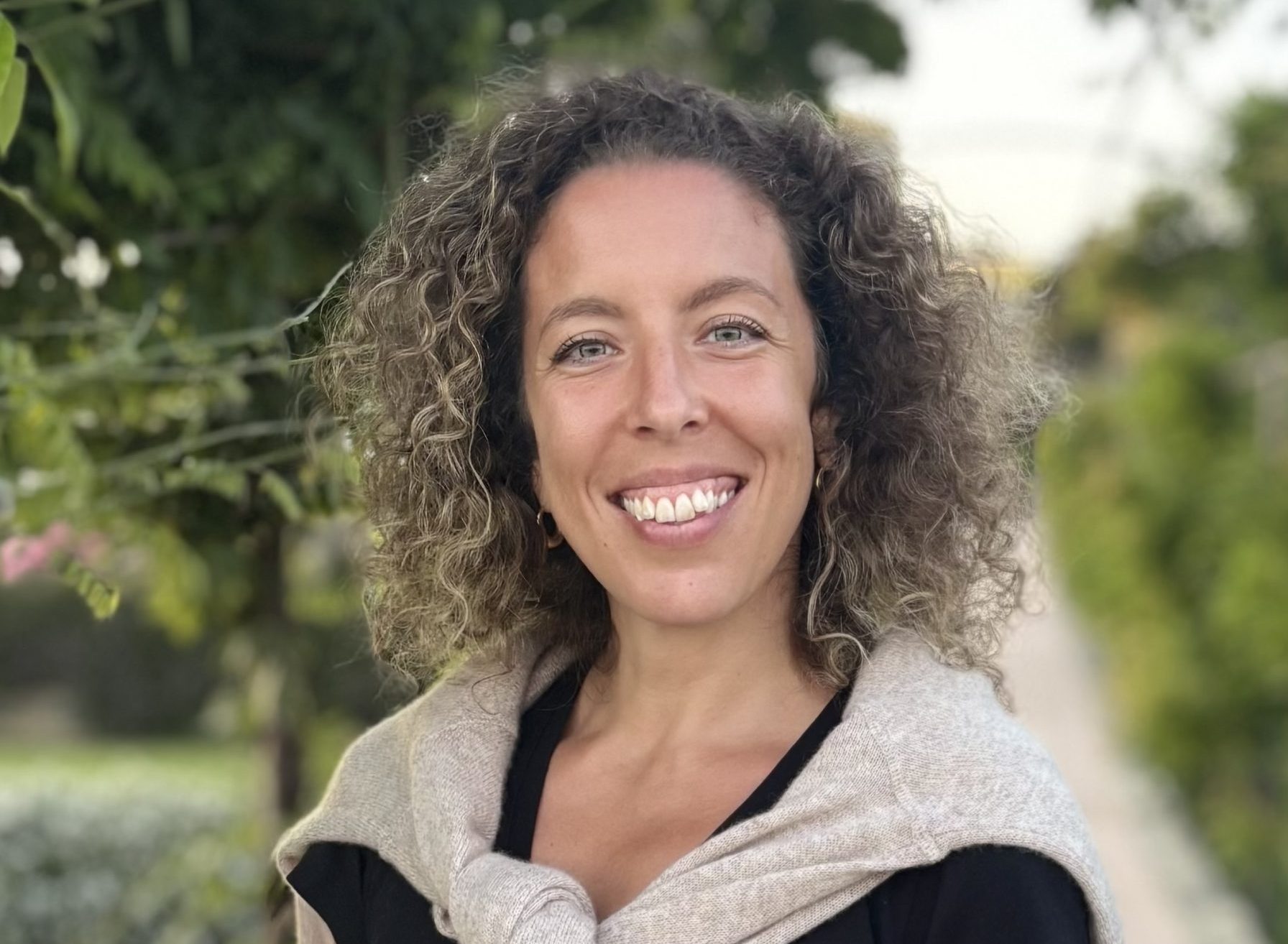 Laura is a fair skinned woman with curly brown hair. She is wearing a black shirt with a tan sweater and is smiling into the camera.